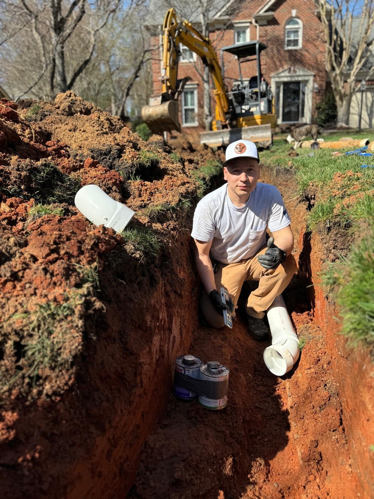 Construction worker crouching in excavated trench with pvc pipe, yellow excavator and brick house in background