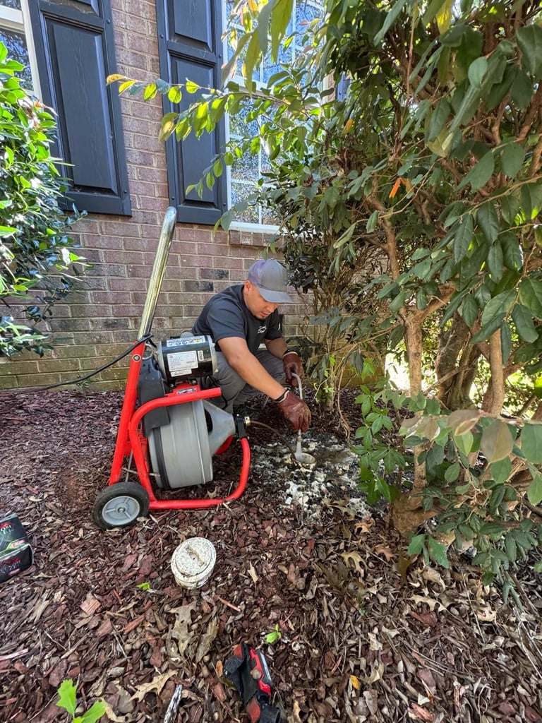 Man operating a red sump pump near brick building with ivy-covered wall