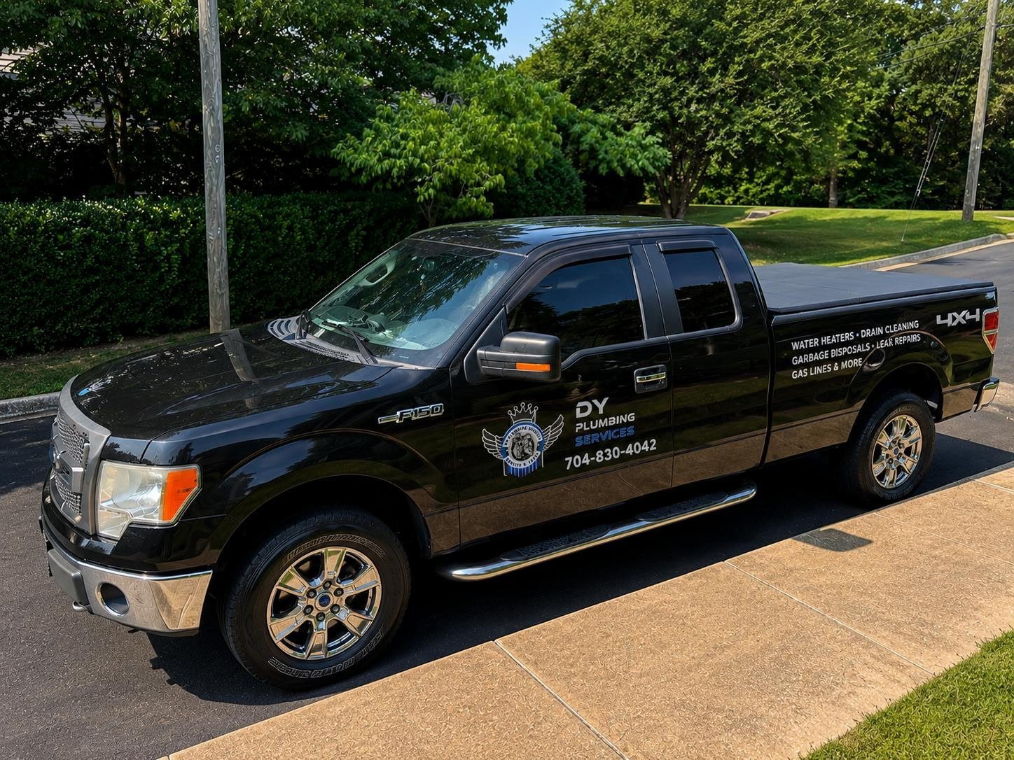 Black Ford F-150 pickup truck with DY Plumbing Services branding parked on a concrete driveway with trees and grass in the background