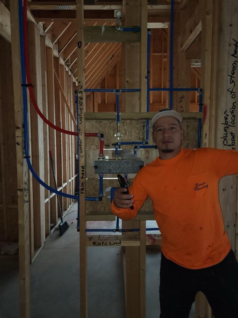 Worker in orange shirt installing blue and red plumbing pipes in residential home construction framed with wood studs