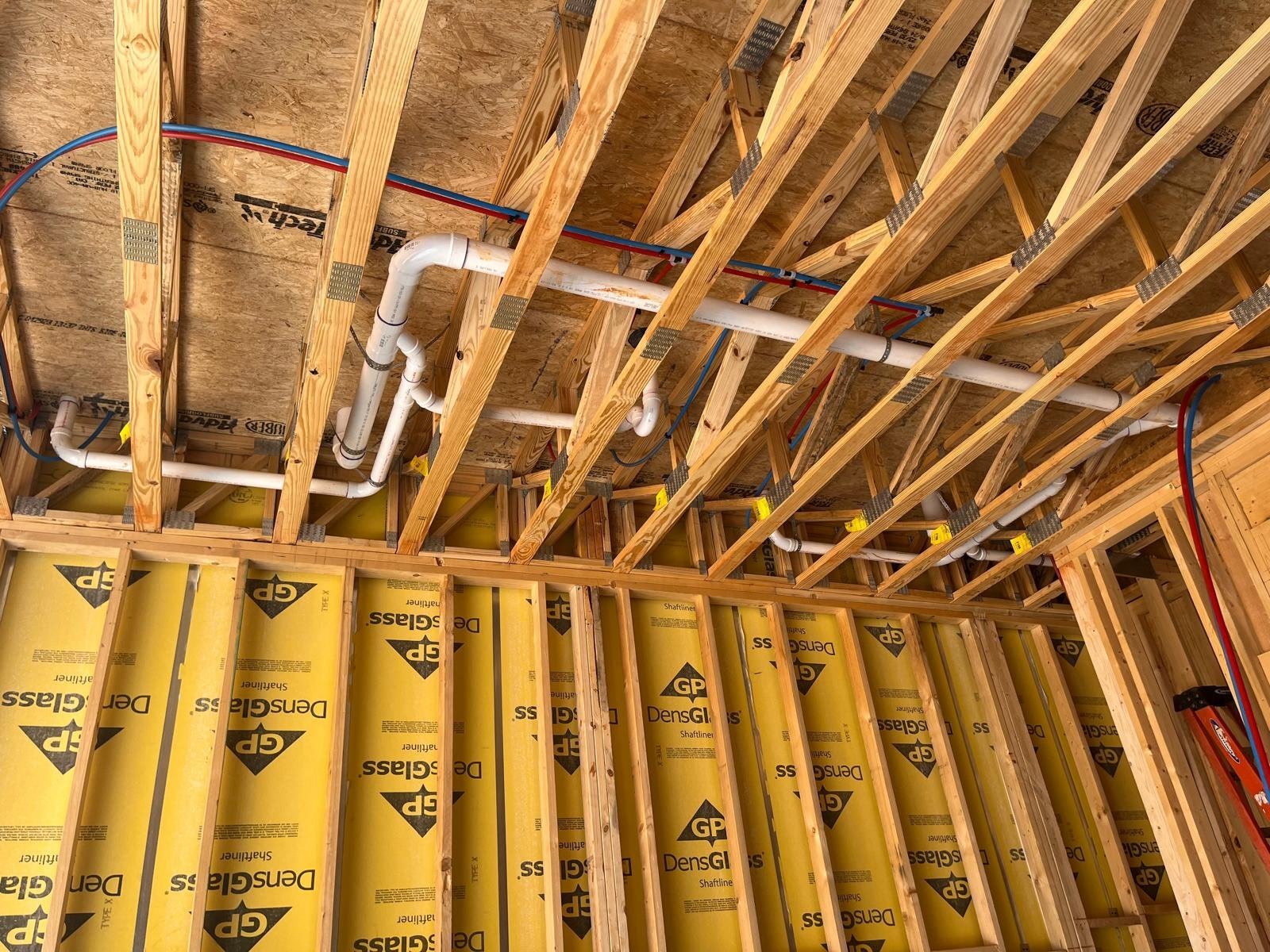Interior view of building framing showing wooden studs, yellow insulation, and white PVC pipes installed in wall cavity during construction.