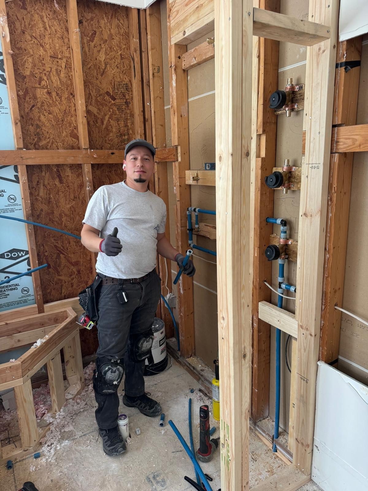Worker in construction site with wooden framing, wearing gray shirt and tool belt, standing near plumbing installation materials